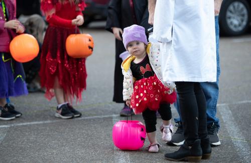 Young child dressed up like minnie mouse peaking out from behind an adult's leg, halloween pumkin baskets in the background